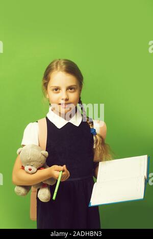 Girl with braids and serious face. Study and back to school concept. Pupil holds blue book, marker and teddy bear. Kid in school uniform isolated on green background Stock Photo