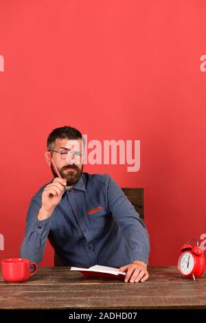 Man with sleepy face sits at wooden table. Cup, retro clock and red book on vintage table. Teacher with beard and glasses holds marker and notebook, red background. Exam and studying concept Stock Photo