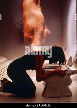 Bulimia. Young woman kneels at a toilet whilst forcing herself to vomit. A hand with downwardly pointing thumb may abstractly depict the force with wh Stock Photo