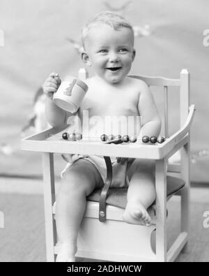 1920S SMILING BABY SITTING IN HIGH CHAIR HOLDING DRINKING FROM BUNNY MUG - b5006 HAR001 HARS BLACK AND WHITE CAUCASIAN ETHNICITY CLOTH DIAPER HAR001 OLD FASHIONED Stock Photo