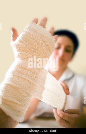 MODEL RELEASED. Treatment after surgery. Nurse applying a bandage to a patient's hand during a home visit. He has had an operation on his hand to corr Stock Photo