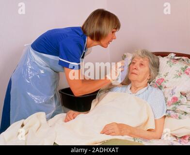 Bed bath. A district nurse gives an elderly bedridden woman a bed bath. The nurse is washing and drying the face of the patient. She will wash a small Stock Photo