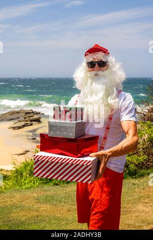 Santa Claus holds gift boxes with the ocean on backgraund. Australia, Christmas in summer time. Stock Photo