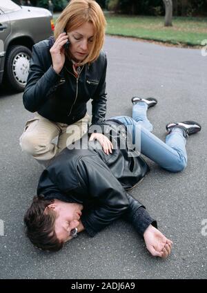 Road accident first aid. Woman using a mobile phone to call for emergency aid for the victim of a road traffic accident (RTA). She has already placed Stock Photo