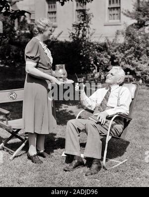 1940s  ELDERLY  COUPLE IN BACKYARD WOMAN SERVING BEVERAGE TO MAN SITTING IN LAWN CHAIR HOLDING GLASS AND BOOK WEARING NECKTIE  - s7722 HAR001 HARS COMMUNICATION LIFESTYLE SATISFACTION ELDER FEMALES MARRIED SPOUSE HUSBANDS HOME LIFE COPY SPACE FRIENDSHIP FULL-LENGTH LADIES PERSONS MALES SENIOR MAN EYEGLASSES SENIOR ADULT B&W PARTNER SENIOR WOMAN PITCHER HAPPINESS OLD AGE OLDSTERS OLDSTER BEVERAGE LEISURE AND HAIRSTYLE NECKTIE ELDERS SUSPENDERS FINGER WAVES CONNECTION DOTTED SWISS ELDERLY MAN COOPERATION ELDERLY WOMAN GRAY GRAY HAIR ICED TEA RELAXATION TOGETHERNESS WIVES BLACK AND WHITE Stock Photo