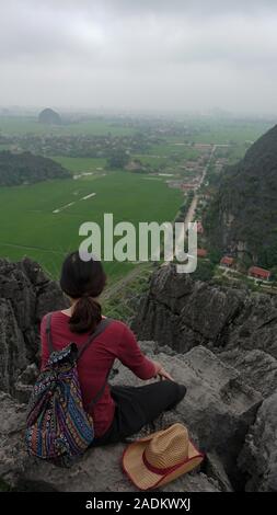 A woman rests, relaxes and enjoys the view of rice fields below after hiking up a mountain near Tam Coc, Ninh Binh, Vietnam Stock Photo