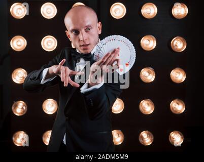 Dexterity of hands. Magician in black suit and with playing cards standing in the room with special lighting at backstage Stock Photo
