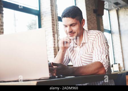 Reading some good news from smartphone. Adult man sits in cafe at daytime and using the laptop for the remote work Stock Photo