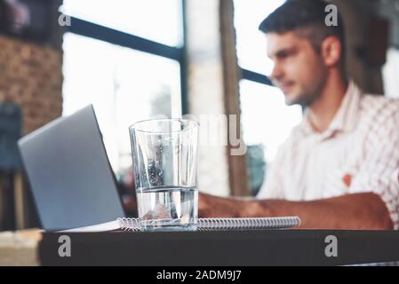 Glass of water in focus. Adult man sits in cafe at daytime and using the laptop for the remote work Stock Photo