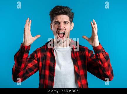 Young european stressed man in red shirt shouting isolated over blue background. Stressed and depressed guy loudly screaming to camera. Stock Photo