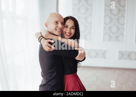 Cute and cheerful people. Hug each other and smiling. Portrait of happy couple indoors. Bald guy and brunette woman stands in the white room Stock Photo