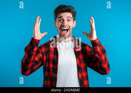 Happy european man rejoices. Handsome guy with stylish hairdo surprised to camera over blue background. Stock Photo