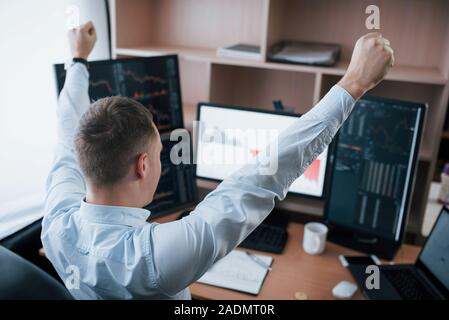 Hands up because of success. Man working online in the office with multiple computer screens in index charts Stock Photo