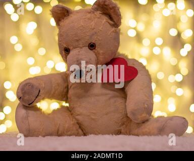 Very old soft toy. Between 40 to 50 years old teddy bear with red paper heart. On the backdrop are christmas lights. Nostalgia, love. Stock Photo