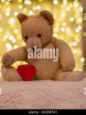 Very old soft toy. Between 40 to 50 years old teddy bear with red paper heart. On the backdrop are christmas lights. Nostalgia, love. Stock Photo