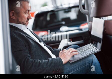 Have job even on the road. Working on a back of car using silver colored laptop. Senior businessman Stock Photo