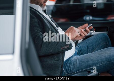Cropped photo. Working on a back of car using silver colored smartphone. Senior businessman Stock Photo