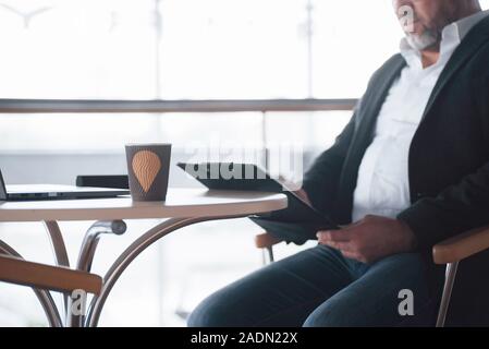 No time for rest, job is not waiting. Boss sits on the chair near the table with silver colored laptop and reads information from notepad Stock Photo