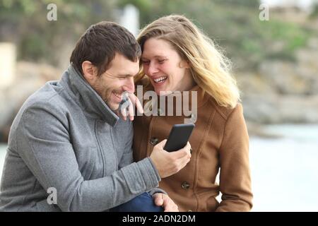 Happy couple laughing loud talking about mobile phone contenton the beach in winter holidays Stock Photo