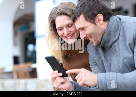 Happy couple of adults checking smart phone online content in the street in winter Stock Photo