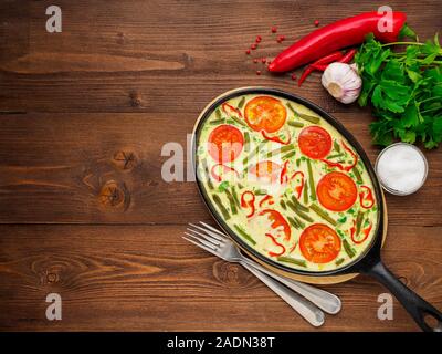 Pan with traditional Italian Frittata on the side of a brown wooden table, forks, parsley, salt, pepper, tomatoes. Top view, empty space for text Stock Photo