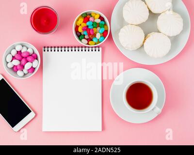 Cup of tea, notepad, mobile and plates of different sweets - marshmallows, lollipops, candy. Pink background, top view, flat lay Stock Photo