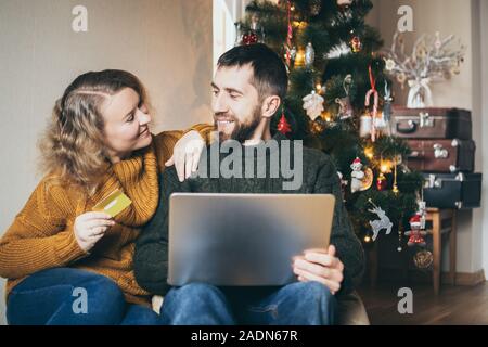 Young Caucasian couple doing Christmas shoppings online on the couch at home with laptop, looking at each other and smiling