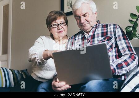 Elderly Caucasian couple doing shoppings online on the couch at home, looking at laptop screen and smiling Stock Photo