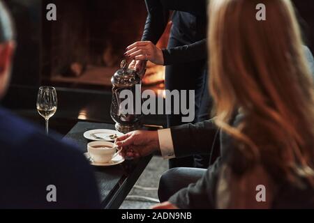 Female waiter pouring hot tea in the white cup. Friends sits in the restaurant with beautiful fireplace in it Stock Photo