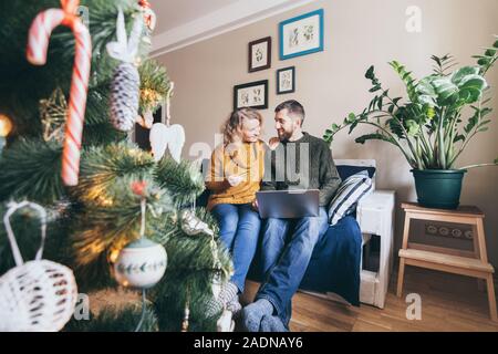 Young Caucasian couple doing Christmas shoppings online on the couch at home with laptop, looking at each other and smiling Stock Photo
