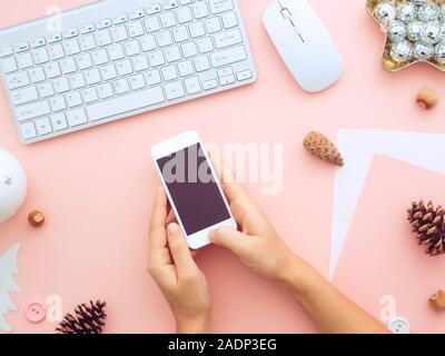 Writing greetings by sms. Christmas female hands holding mobile phone on a desk with keyboard, and New Year decorations on it. Feminine blogger winter Stock Photo