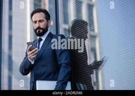 Serious bearded businessman leaning against glass wall Stock Photo