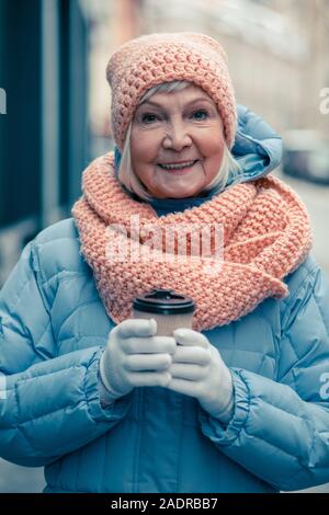 Aged woman looking glad while standing outdoors with coffee Stock Photo