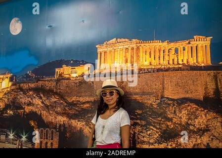 Asian woman in white t-shirt in Athens in white hat, standing in front of large poster of Acropolis with full moon under the Acropolis Stock Photo