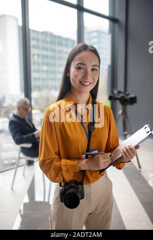 Dark-haired young cute reporter with camera feeling happy Stock Photo