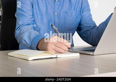 Businessman using laptop, taking notes at office desk - Close up. Man using computer and writing notes to notebook at office desk. Busy business man w Stock Photo