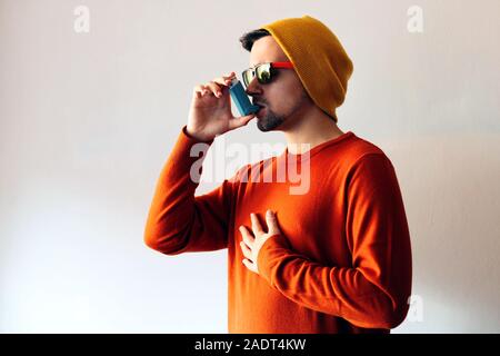 Man dressed in orange and wearing a yellow cap using a blue asthma inhaler to prevent an asthma attack. Medicine and health Concept. Stock Photo