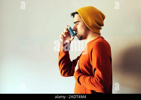 Man dressed in orange and wearing a yellow cap using a blue asthma inhaler to prevent an asthma attack. Medicine and health Concept. Stock Photo
