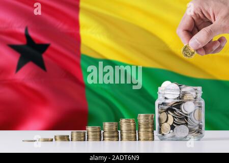 Business man holding coins putting in glass, Guinea Bissau flag waving in the background. Finance and business concept. Saving money. Stock Photo