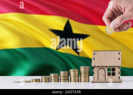 Man holding coins putting in wooden house moneybox, Ghana flag waving in the background. Saving money for mortgage. Stock Photo