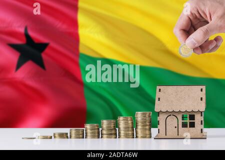 Man holding coins putting in wooden house moneybox, Guinea Bissau flag waving in the background. Saving money for mortgage. Stock Photo