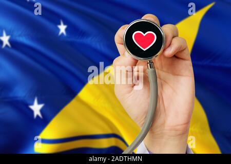 Doctor holding stethoscope with red love heart. National Tokelau flag background. Healthcare system concept, medical theme. Stock Photo