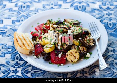 Grilled vegetables salad with feta cheese in white plate on blue textile background. Close up Stock Photo