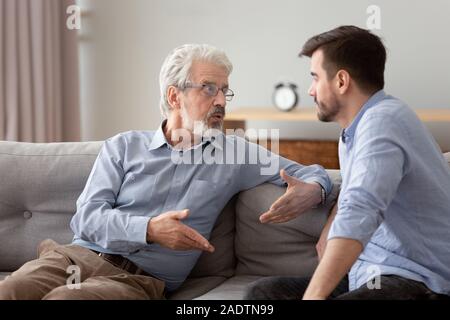 Serious elderly father and grownup son sitting on couch talking Stock Photo