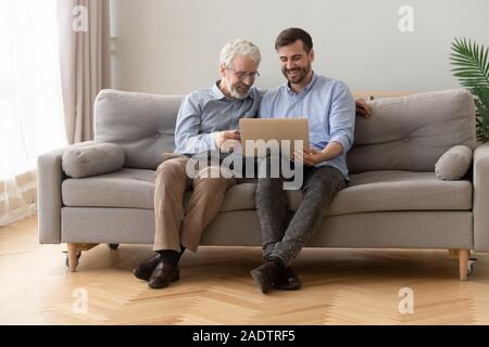 Father and grown up son sit together on sofa with computer Stock Photo