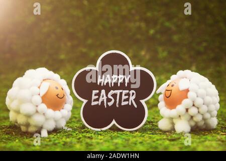 funny lamb figures and flower shaped slate with english text happy easter, green background with sunshine Stock Photo