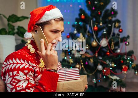 Left alone for Christmas. Sad man talking to friends on smartphone holding gift boxes. Celebrating New year at home. Stock Photo