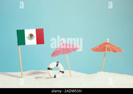 Miniature flag of Mexico on beach with colorful umbrellas and life preserver. Travel concept, summer theme. Stock Photo