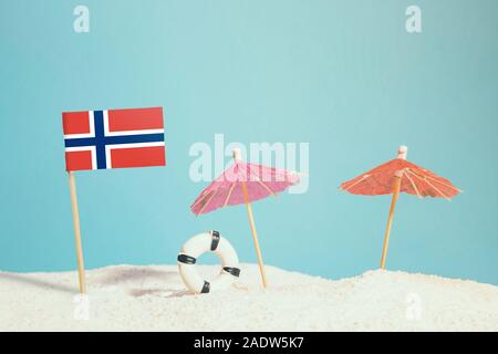 Miniature flag of Norway on beach with colorful umbrellas and life preserver. Travel concept, summer theme. Stock Photo