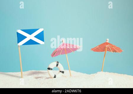 Miniature flag of Scotland on beach with colorful umbrellas and life preserver. Travel concept, summer theme. Stock Photo